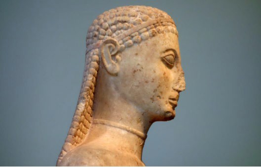 Side view of the head of a kouros. The hair is plaited, and the eyes sit on the surface of the face. The ears are large and resemble those on Egyptian statuary. There is a band around the neck.