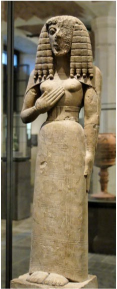 A carved stone statue of a woman. She stands stiffly with plaited hair. Half of her face is no longer present. Her fingers are overly long and delicate.