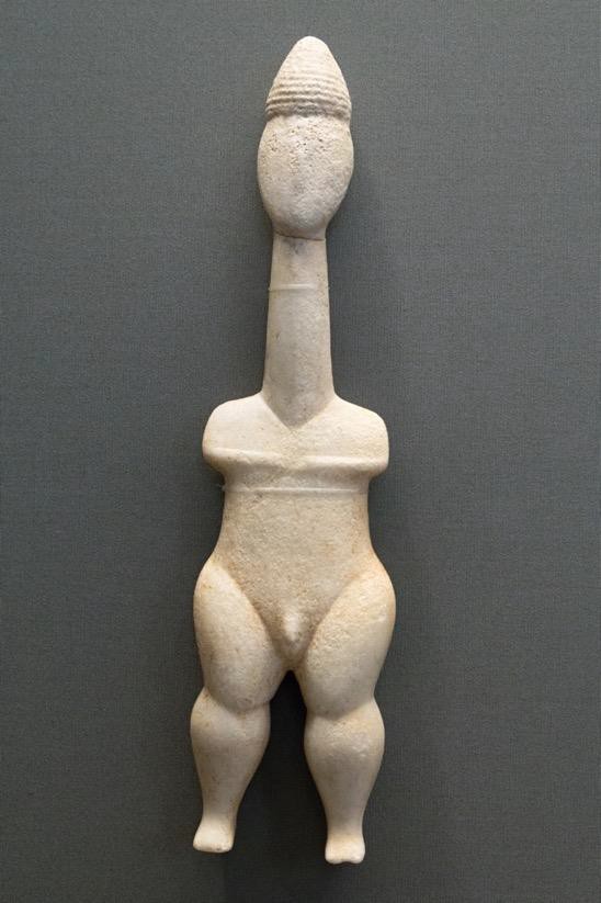 A simplified white stone figure of a male. It is stylized with a long neck and small torso. This contrasts with strong legs.