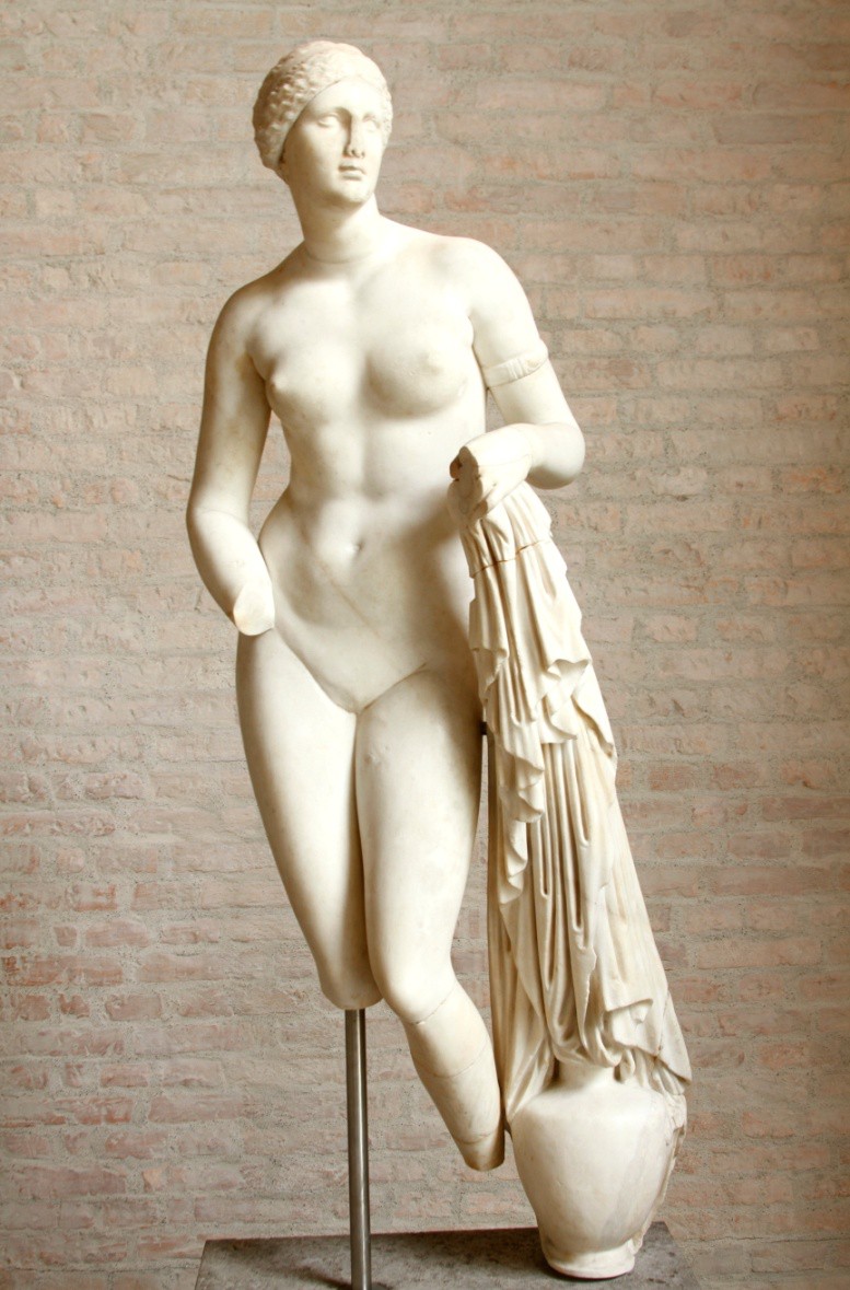 A marble statue of a nude Aphrodite. She holds a cloth in her right hand that trails across a pottery vessel and her other hand, now missing, was held modestly in front of her groin.