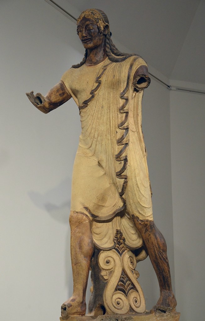 A large terra cotta statue of Apollo that is more natural than an Archaic styled statue but does have one foot forward and a smile. The statue is clothed. His eyes sit on the surface of his face like those of an Archaic Greek.