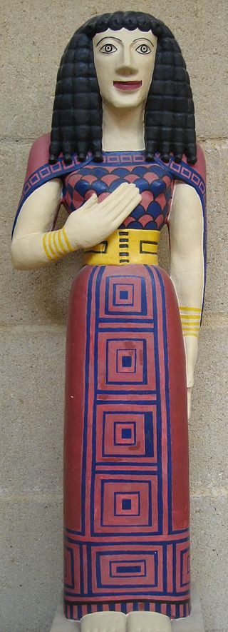 A painted replica of Lady of Auxerre. She is brightly painted with a geometrically decorated dress that reaches her feet and covers her shoulders. She has bracelets on each wrist.