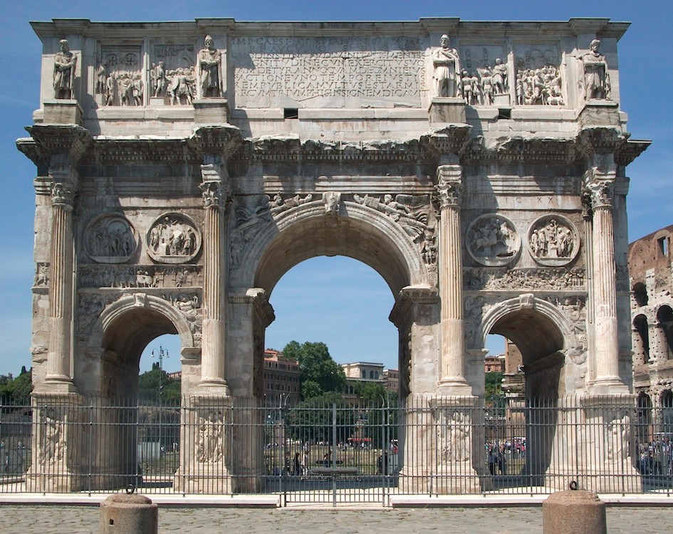 A carved Roman triumphal arch with a fence around it. It has a large arch in the center with a smaller arch on each side of it. There are attached columns and medallions. Statues and two bas reliefs top the monument. There is writing in the center of the top section, over the central arch.