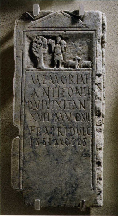 A stele with a carving of Christ the shepherd carrying a sheep over his shoulders. He stands near a tree and other sheep. There is writing below the image.