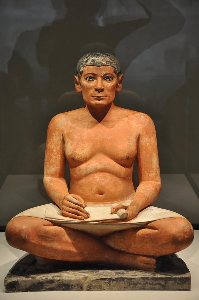 A small but otherwise realistic stone carving of a seated scribe. He holds his paper and pen on his lap.