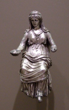 A silver statue of a woman who appears to hold an offering in her right hand. She is fully clothed in long robes and is seated.