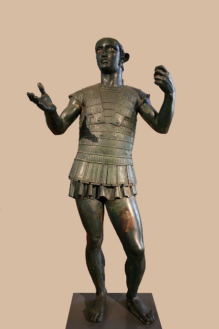 A bronze statue of a young man in battle gear His helmet is missing. His eyes are inset as they came to be during the Greek classical age.