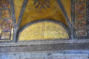 A golden mosaic with the outline of a cross in it. On each side of that arch are flower-like mosaics in bands.